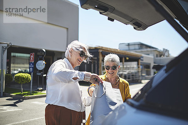 Ältere Freundinnen laden Einkaufstüten auf einem sonnigen Parkplatz in den Kofferraum eines Autos