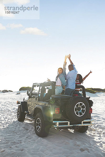 Übermütige junge Freunde  die mit erhobenen Armen im Jeep am Strand jubeln und einen Road Trip genießen