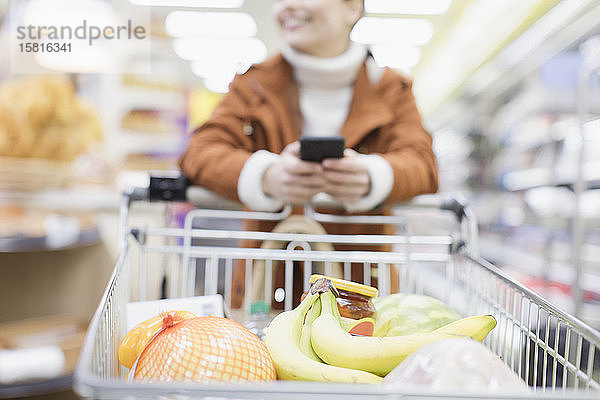 Frau mit Smartphone schiebt Einkaufswagen im Supermarkt