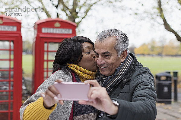 Älteres Paar  das sich küsst und ein Selfie im Park vor einer roten Telefonzelle macht