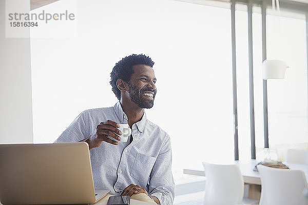 Lächelnder  begeisterter Mann  der Kaffee trinkt und zu Hause am Laptop arbeitet