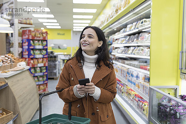 Lächelnde junge Frau mit Smartphone beim Lebensmitteleinkauf im Supermarkt