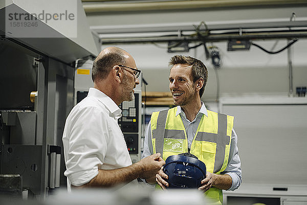 Geschäftsmann im Gespräch mit lächelndem Mann in reflektierender Weste in einer Fabrik