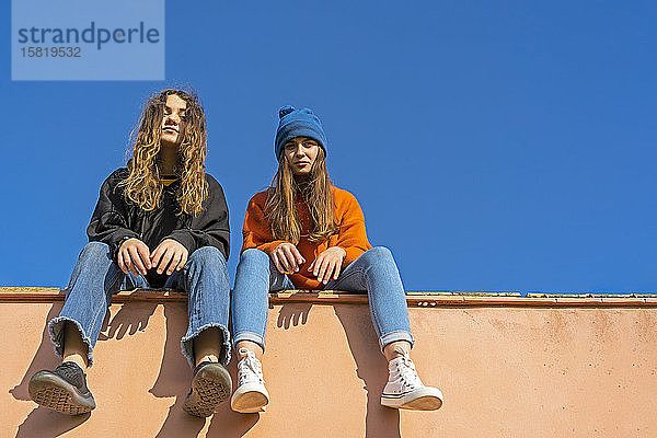 Porträt von Mädchen im Teenageralter  die an einer Wand vor blauem Himmel sitzen