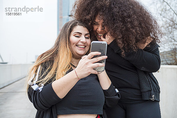 Zwei glückliche  sportliche junge Frauen  die in der Stadt ein Smartphone benutzen