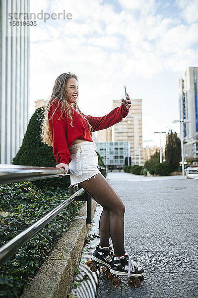 Lächelnde junge Frau auf Rollschuhen beim Selfie in der Stadt
