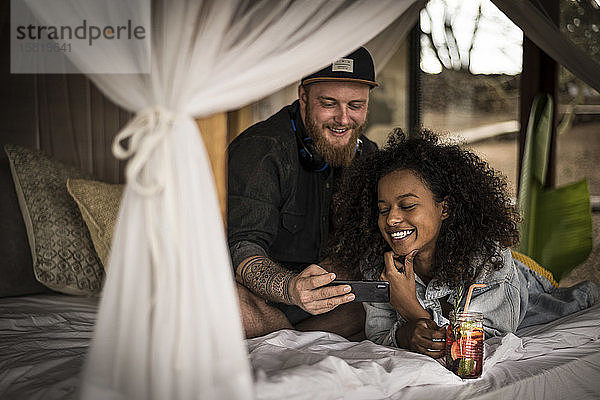 Glückliches Paar im Bett in einer Lodge mit Smartphone und frischem Eistee