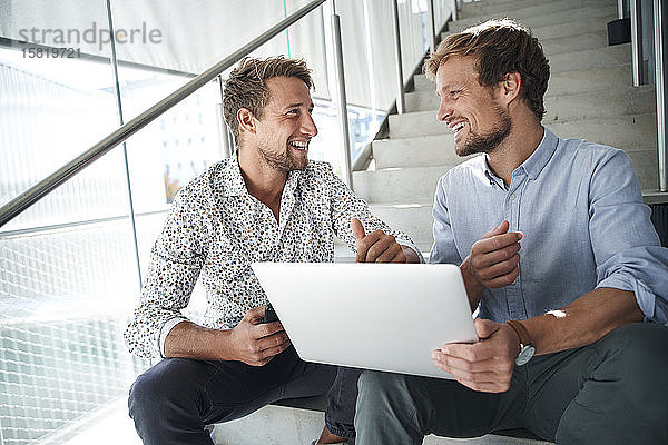 Zwei glückliche junge Geschäftsleute sitzen mit Laptop auf der Treppe