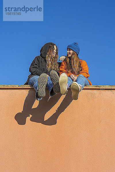 Zwei lächelnde Teenager-Mädchen sitzen auf einer Mauer vor blauem Himmel