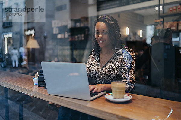 Junge Frau mit einer Kamera mit Laptop in einem Café hinter einer Fensterscheibe