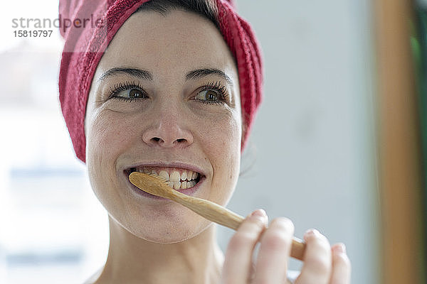 Porträt einer Frau mit in ein Handtuch gewickeltem Kopf beim Zähneputzen mit einer hölzernen Zahnbürste