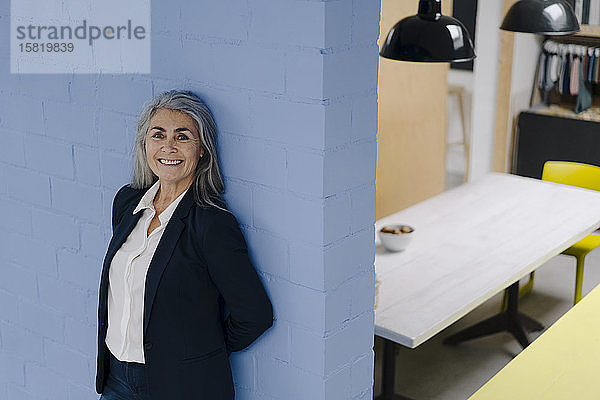 Porträt einer lächelnden grauhaarigen Geschäftsfrau  die an einer blauen Wand in einem Loft-Büro steht