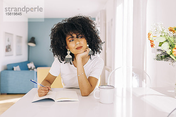 Porträt einer jungen Frau mit lockigem Haar  die zu Hause mit einem Notizbuch an einem Tisch sitzt