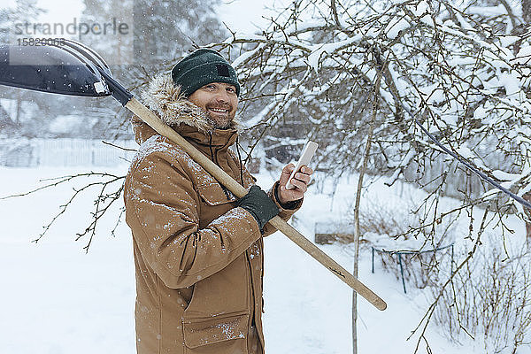 Porträt eines lächelnden Mannes mit Schneeschaufel und Mobiltelefon