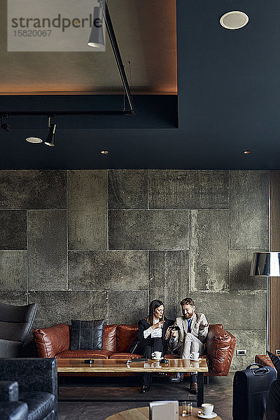 Geschäftsmann und Geschäftsfrau sitzen auf der Couch in der Hotellobby und teilen sich ein Smartphone