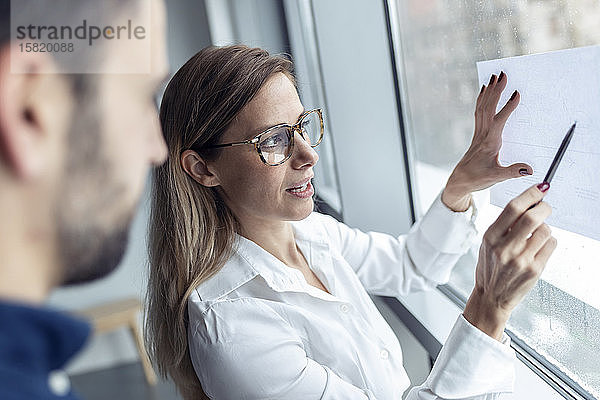 Geschäftsfrau hält ein Blatt Papier am Fenster und zeichnet ein Diagramm  während ein Kollege zusieht