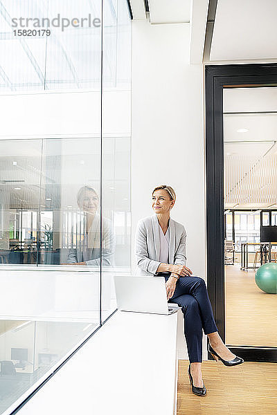 Blonde Geschäftsfrau mit Laptop  auf der Fensterbank eines Bürogebäudes sitzend