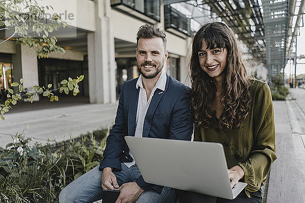 Porträt eines lächelnden Geschäftsmannes und einer lässigen Geschäftsfrau  die einen Laptop im Freien benutzen