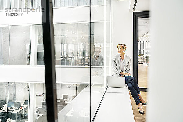 Blonde Geschäftsfrau mit Laptop  auf der Fensterbank eines Bürogebäudes sitzend