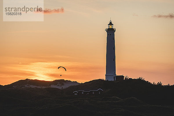Dänemark  Hvide Sande  Leuchtturm vor launischem Himmel in der Dämmerung mit der Silhouette eines Gleitschirms im Hintergrund