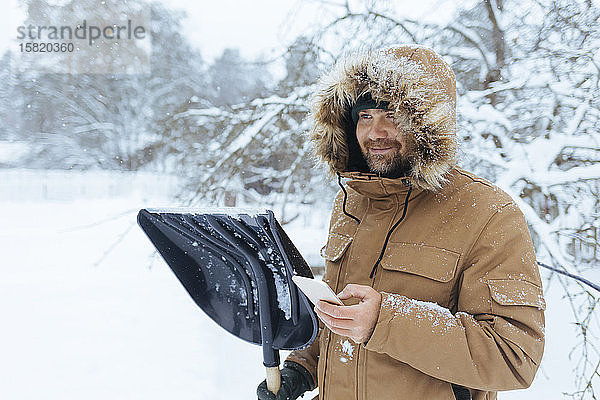 Porträt eines lächelnden Mannes mit Schneeschaufel und Mobiltelefon