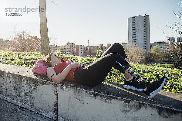Sportliche junge Frau mit Beinprothese  die an einer Wand liegt und telefoniert