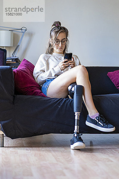 Junge Frau mit Beinprothese sitzt zu Hause auf dem Sofa und benutzt ein Smartphone