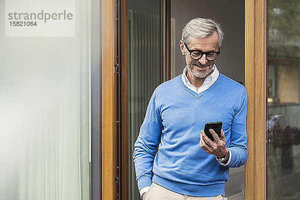 Lächelnder älterer Mann mit grauen Haaren steht vor seinem modernen Design-Haus und schaut auf sein Smartphone