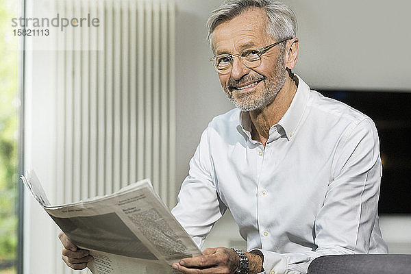 Porträt eines lächelnden älteren Mannes mit grauen Haaren im modern gestalteten Wohnzimmer beim Zeitungslesen
