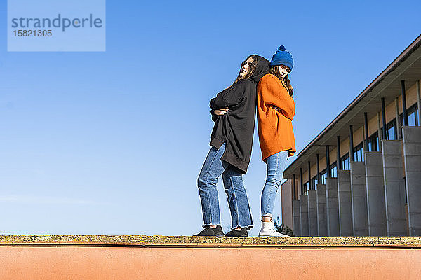 Zwei Teenager-Mädchen stehen Rücken an Rücken an einer Wand vor blauem Himmel