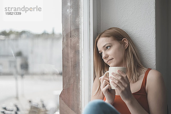 Porträt einer nachdenklichen jungen Frau mit einer Tasse Kaffee  die aus dem Fenster schaut