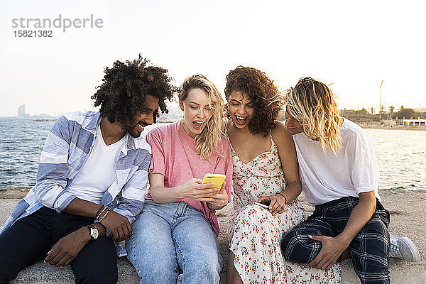 Glückliche Freunde sitzen bei Sonnenuntergang mit Smartphone an der Kaimauer