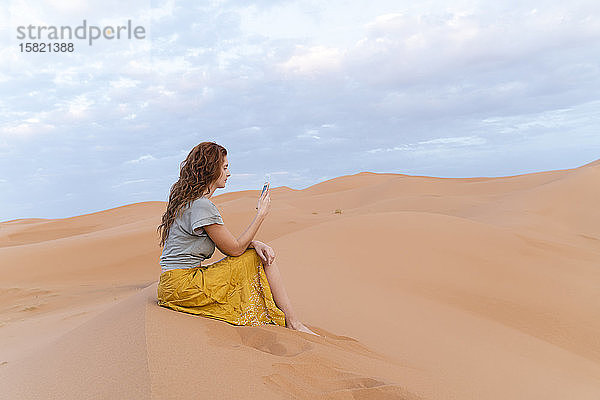 Junge Frau sitzt in einer Sanddüne in der Wüste Sahara und benutzt ihr Handy  Merzouga  Marokko