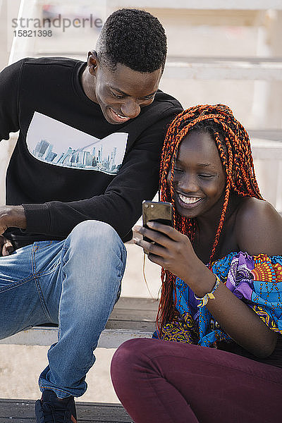 Glückliche Teenagerin und junger Mann sitzen auf der Treppe und schauen auf ihr Handy