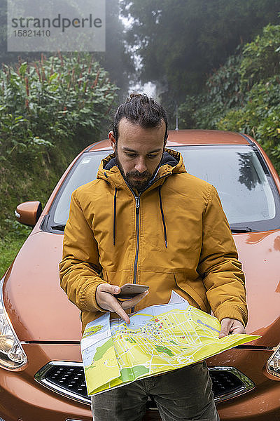 Mann mit Smartphone und Karte vor dem Auto auf Waldstraße  Sao-Miguel-Insel  Azoren  Portugal