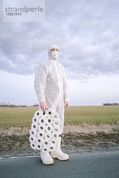 Mann mit Schutzanzug und Maske auf Landstraße stehend mit Toilettenpapier