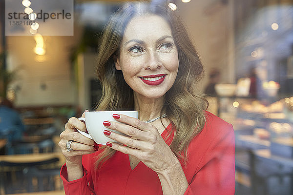 Porträt einer lächelnden reifen Frau mit einer Tasse Kaffee hinter einer Fensterscheibe