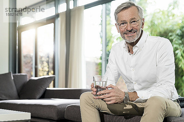 Porträt eines lächelnden älteren Mannes im modern gestalteten Wohnzimmer  der auf einer Couch sitzt und ein Glas Wasser hält
