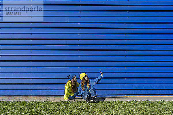 Zwei Mädchen im Teenager-Alter in passender Kleidung nehmen sich mit Smartphone vor blauem Hintergrund ein
