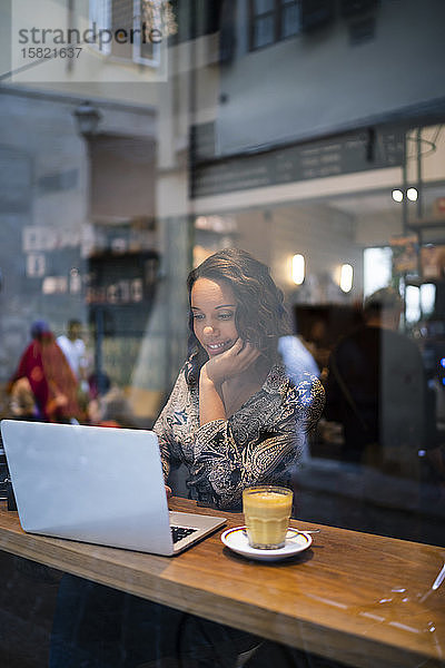 Junge Frau mit Laptop in einem Café hinter einer Fensterscheibe
