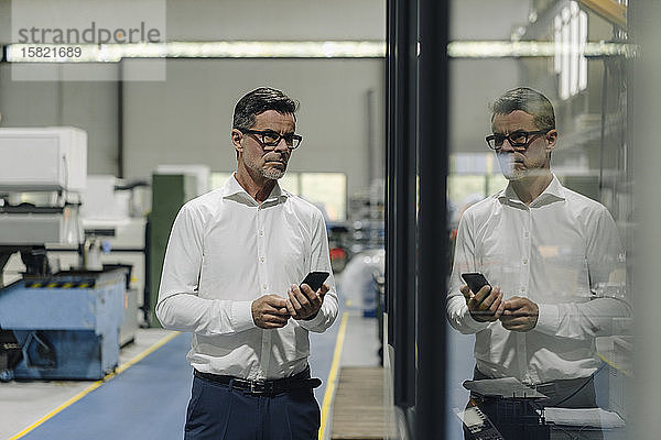 Reifer Geschäftsmann mit Handy in einer Fabrik  das sich in einer Glasscheibe spiegelt