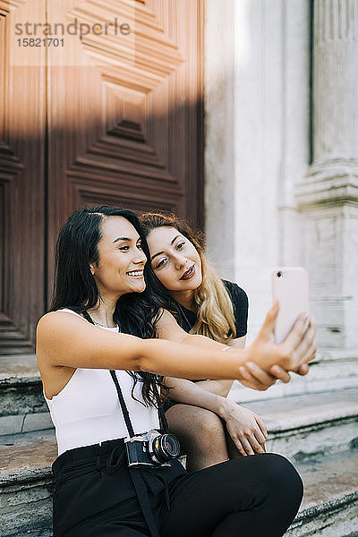 Porträt von zwei Freunden  die auf einer Treppe sitzen und sich mit einem Smartphone selbstständig machen  Lissabon  Portugal