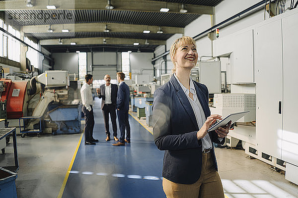 Porträt einer lächelnden Geschäftsfrau mit Tablette in einer Fabrik mit Kollegen im Hintergrund
