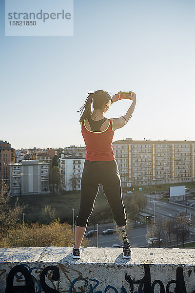 Sportliche junge Frau mit Beinprothese steht an einer Wand über der Stadt und macht ein Smartphone-Foto  Mailand  Italien