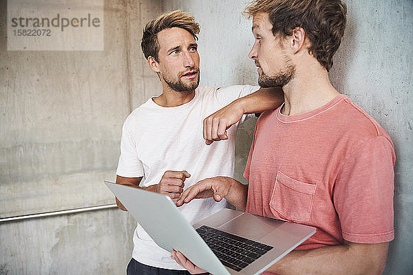 Zwei ungezwungene junge Männer stehen mit einem Laptop an einer Betonwand