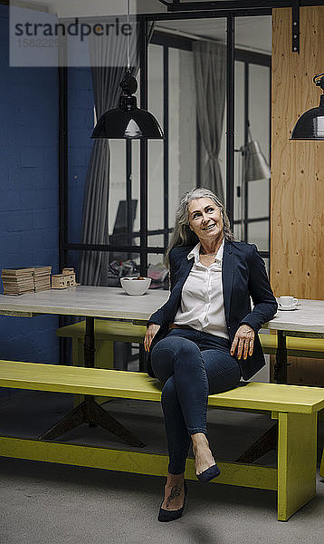 Lächelnde grauhaarige Geschäftsfrau sitzt auf einer Bank in einem Loft-Büro
