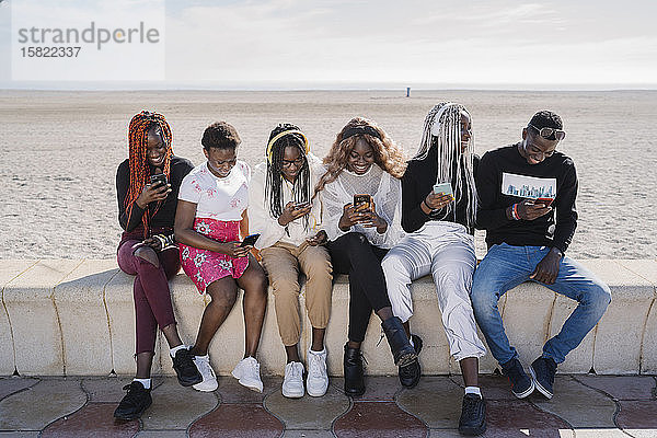 Glückliche Teenager-Freundinnen und junger Mann sitzen mit Smartphones an einer Wand am Strand