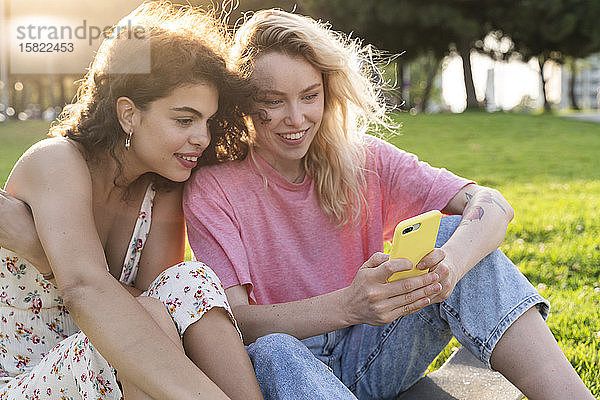 Zwei junge Frauen benutzen ein Smartphone auf einer Wiese im Park
