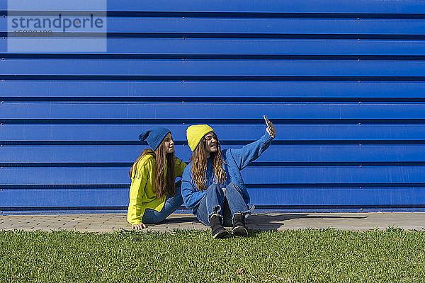 Zwei Mädchen im Teenager-Alter in passender Kleidung nehmen sich mit Smartphone vor blauem Hintergrund ein