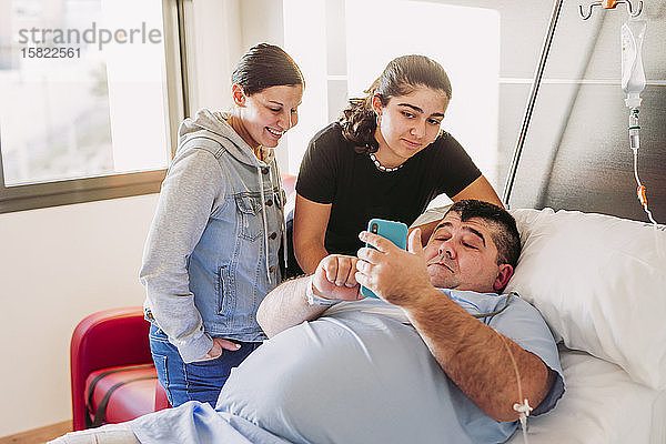 Familie besucht Mann mit Handy im Krankenhausbett liegend
